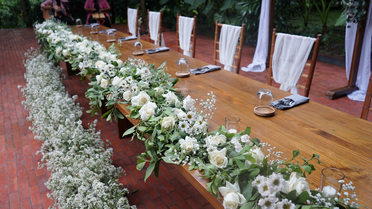 Long wooden wedding reception table decorated with elegant white floral arrangements in a tropical outdoor setting.