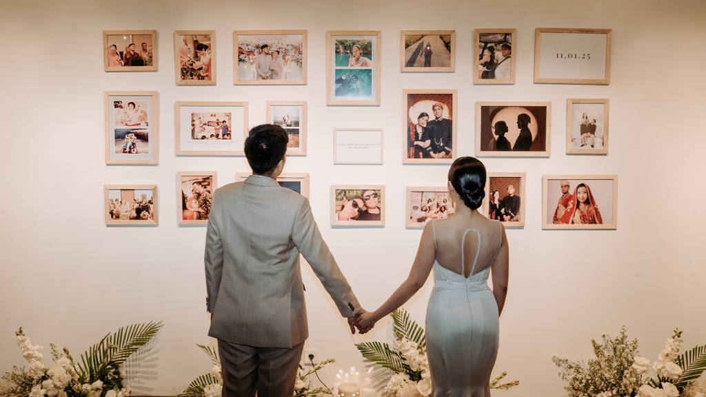 Newlywed couple holding hands while looking at a wall filled with framed wedding photos.