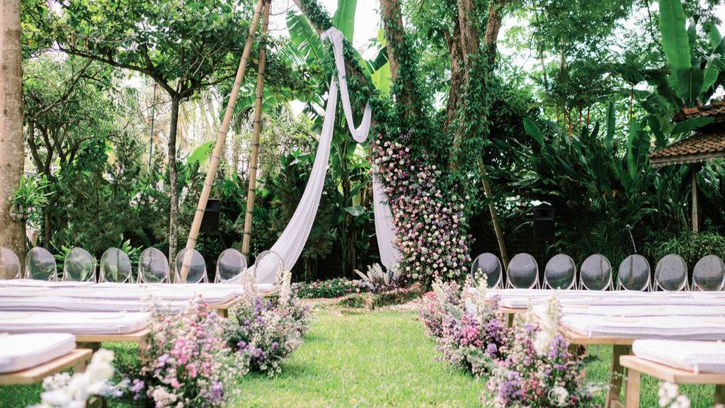 Romantic outdoor wedding ceremony setup with pink and purple floral arrangements, white drapery, and guest seating surrounded by a lush tropical garden.
