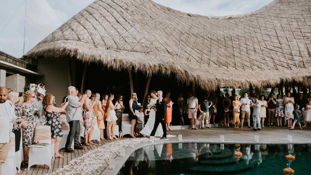 Bride and groom walking beside a swimming pool as wedding guests celebrate at a tropical venue with a large thatched roof.