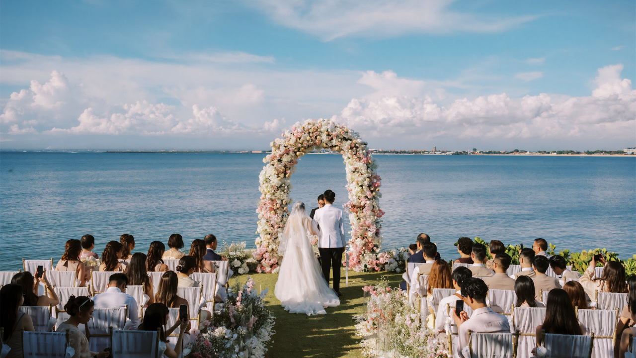 Bride and groom exchanging vows beneath a floral wedding arch overlooking the ocean, surrounded by seated wedding guests.