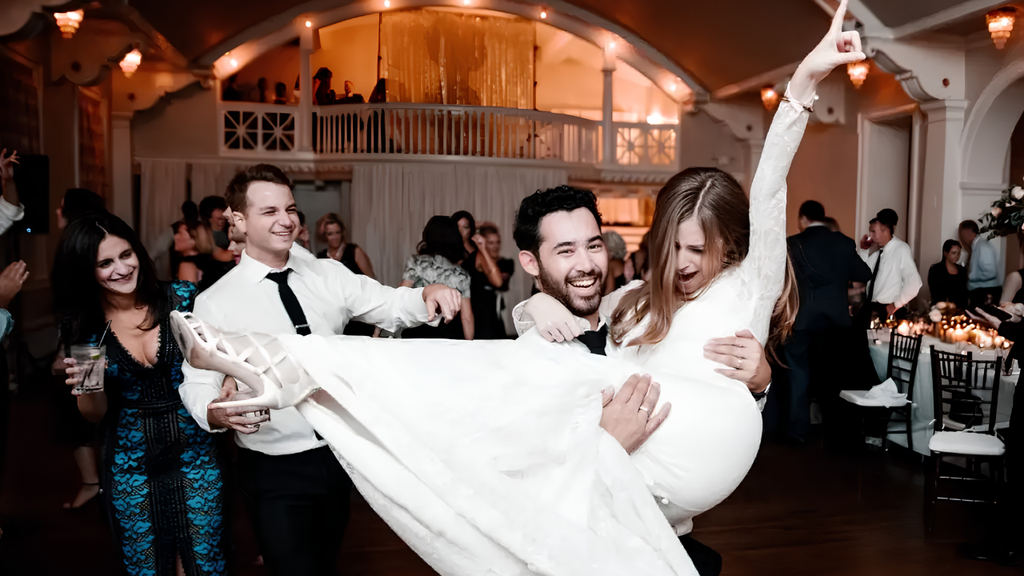 Wedding guests celebrating at an indoor reception as a man lifts a woman in a white dress onto the dance floor.