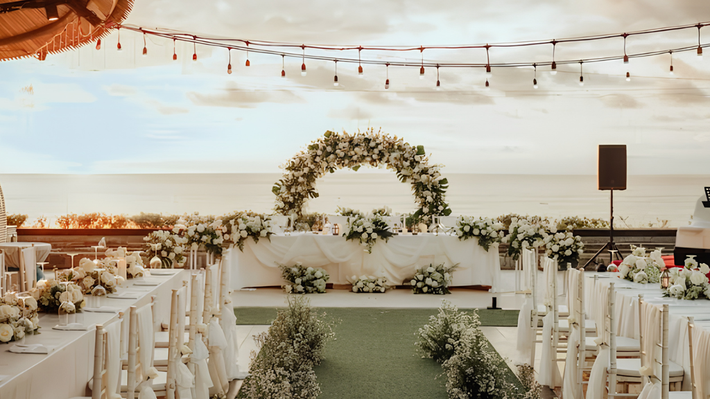 Romantic beachfront wedding setup featuring a white floral arch, elegant dining tables, and ocean views at sunset.