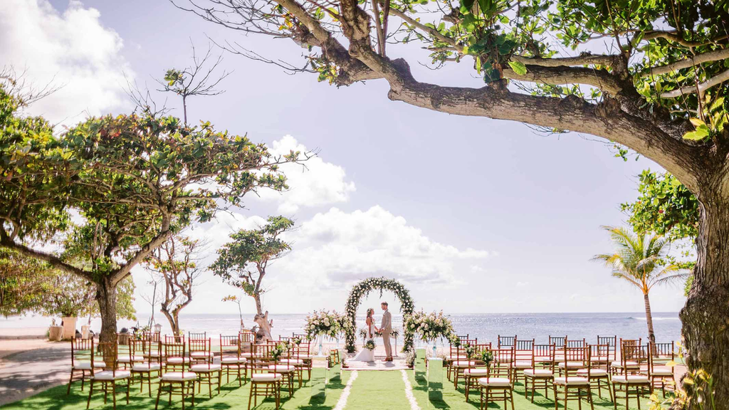 Romantic beachfront wedding ceremony featuring a couple beneath a floral arch, surrounded by wooden chairs and tropical trees.