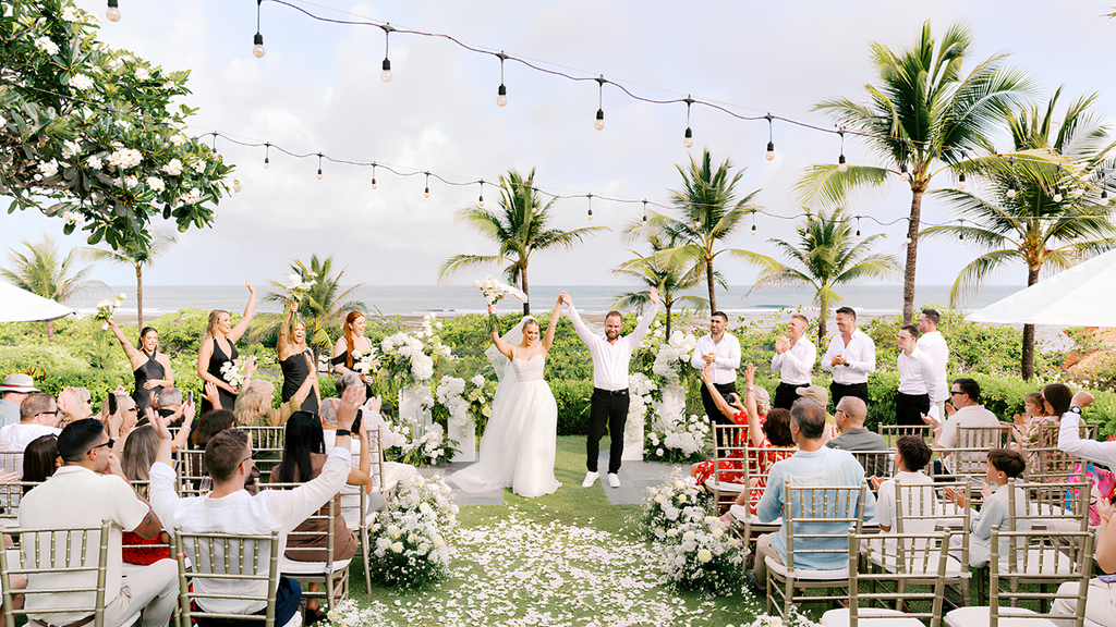 Bride and groom celebrating their wedding ceremony with guests at a tropical beachfront venue decorated with white flowers.