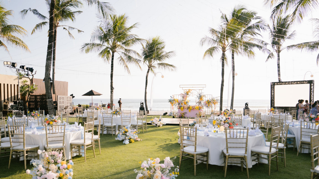 Elegant beachfront wedding reception featuring round dining tables, colorful floral arrangements, palm trees, and ocean views.