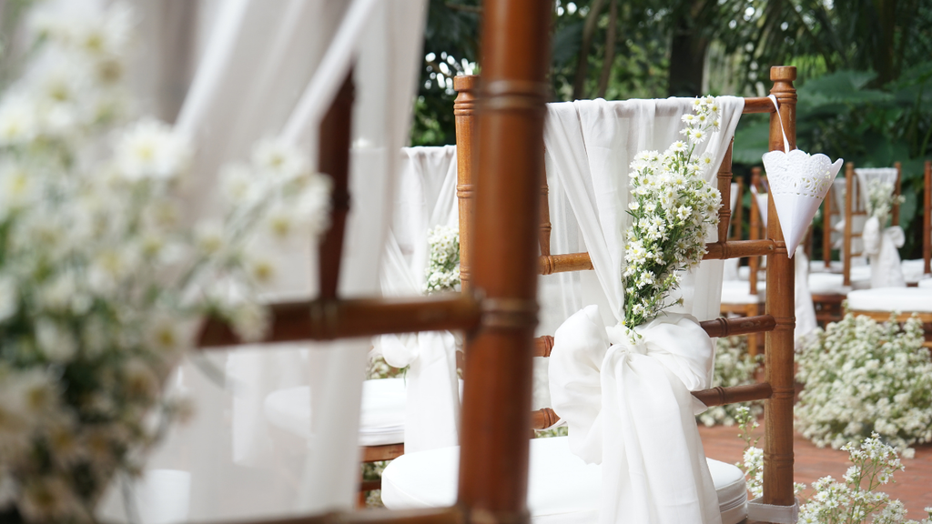 Close-up of wooden wedding chairs decorated with flowing white fabric and delicate white flowers in a tropical garden.