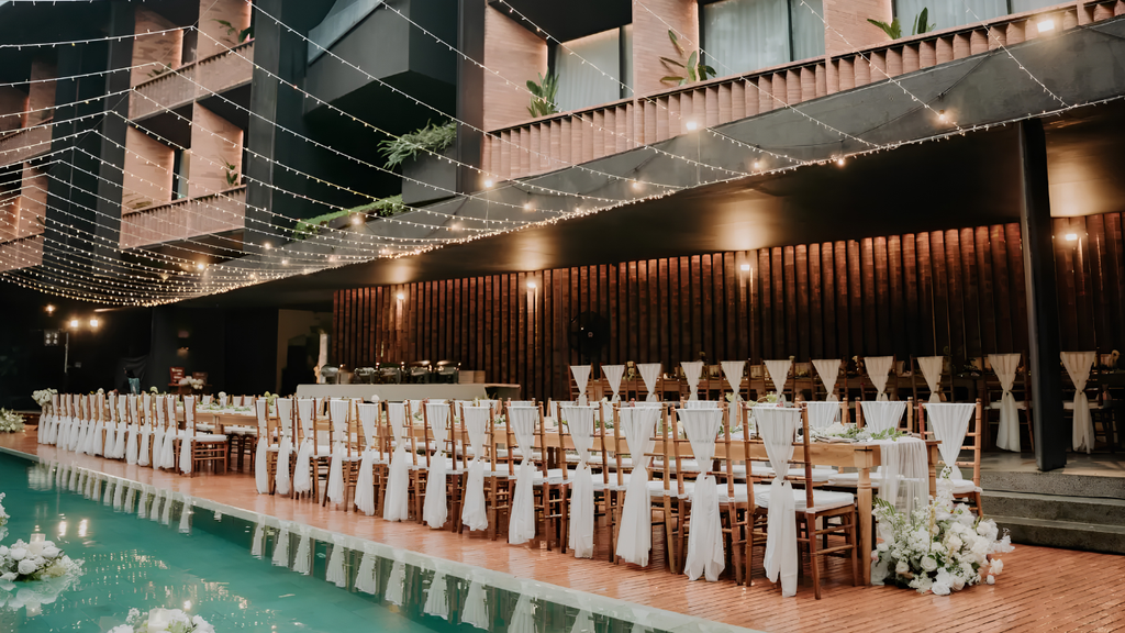Elegant evening wedding reception beside a swimming pool, featuring long dining tables, white floral decorations, and warm string lights.