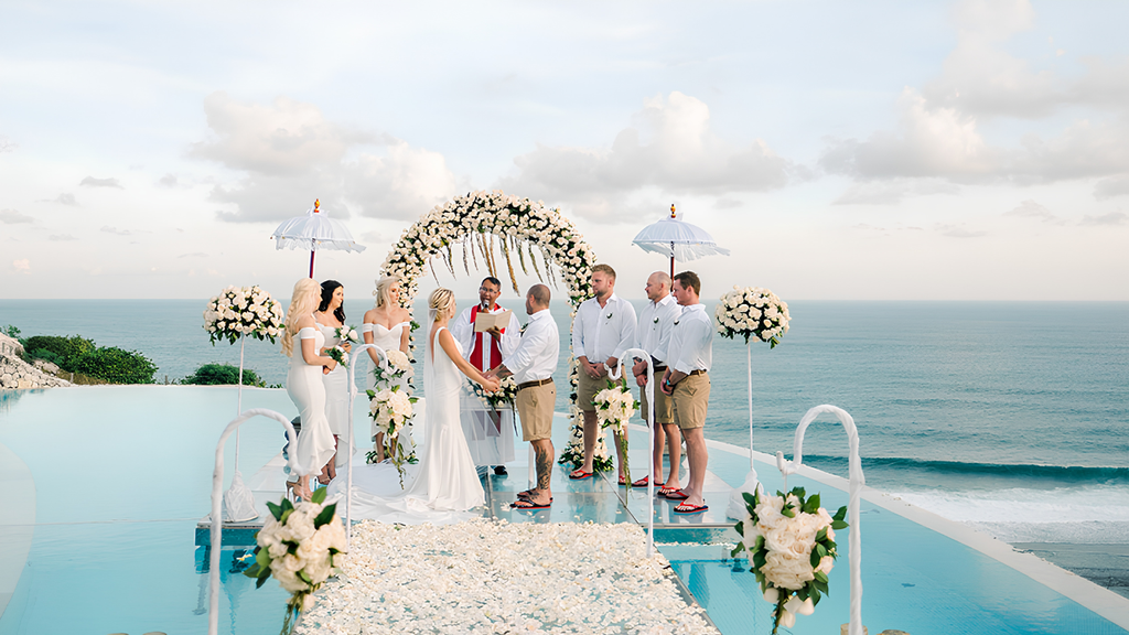 Intimate wedding ceremony on a platform surrounded by an infinity pool, featuring white floral arrangements and panoramic ocean views.
