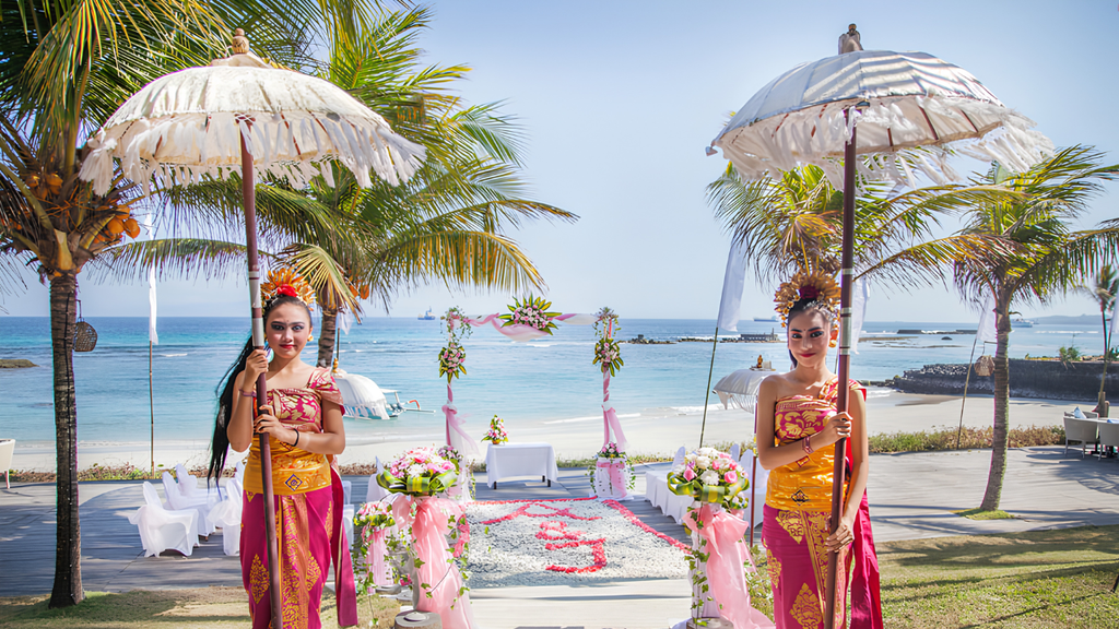 Two women wearing traditional Balinese attire holding ceremonial umbrellas beside a decorated beachfront wedding aisle.